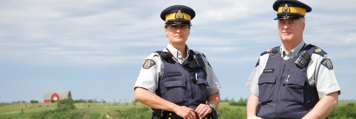 Male and Female RCMP officers stand near a parked police car on a rural road.