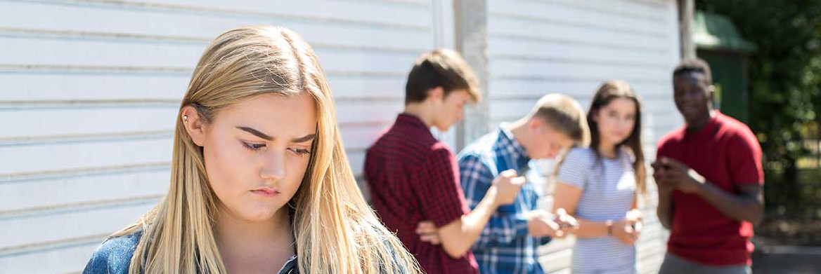 En avant plan, une adolescente tenant un téléphone a l'air triste. À une certaine distance derrière elle, quatre adolescents (trois garçons et une fille) sont debout en train de regarder leur téléphone ou de la fixer.