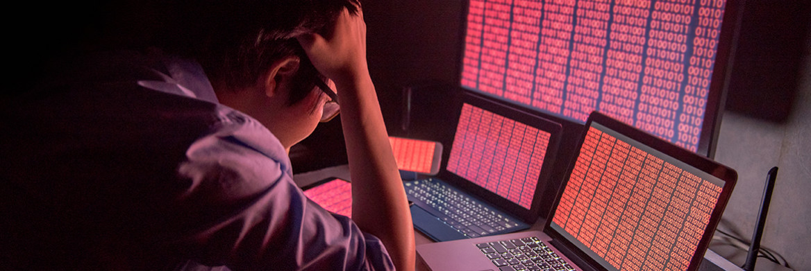 A person, sitting at a desk in front of several computer screens, holds his head in his hands.