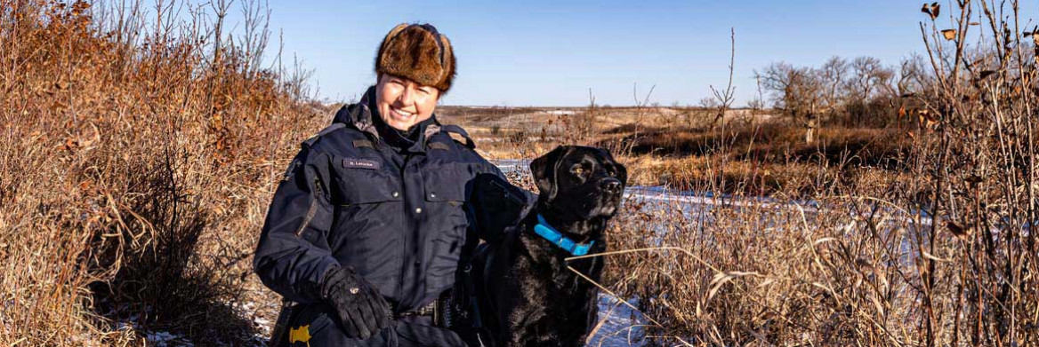 Dans une zone boisée et un décor hivernal, une policière en uniforme se tient accroupie au côté d'un chien noir.
