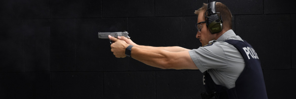 Male officer aims and points a pistol to the left in front of a dark background.