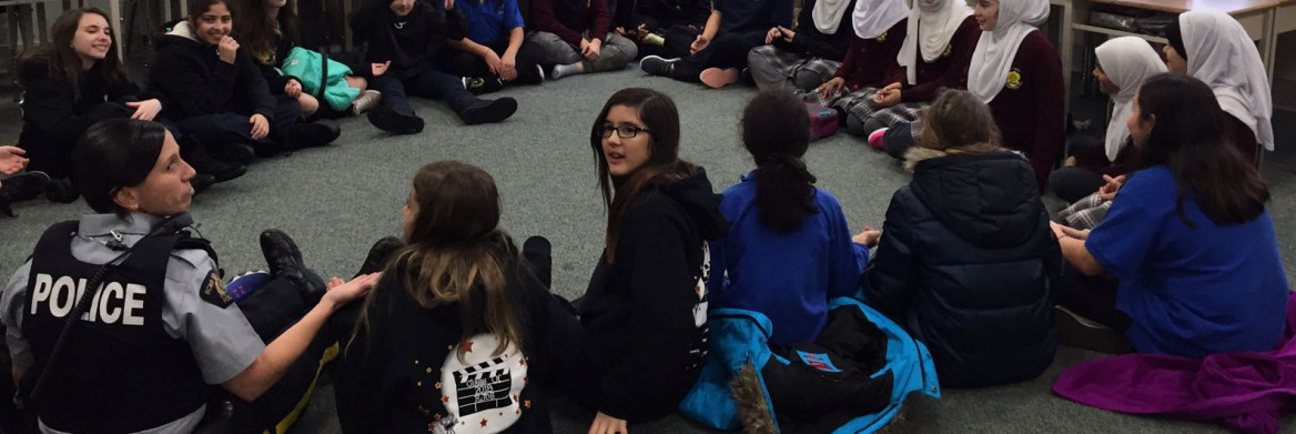 Female police officer sits in a circle on the floor of a classroom with young female students, some of whom are wearing head coverings.