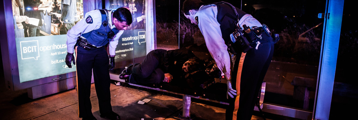 Deux policiers regardent une personne dormir sur le banc d'un abribus la nuit.