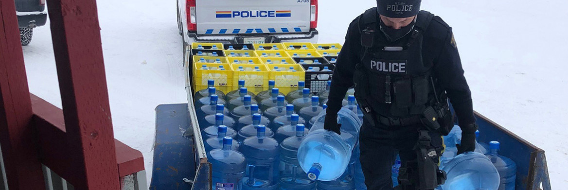 An RCMP officer carries two large water containers. An RCMP truck and trailer are in the background.