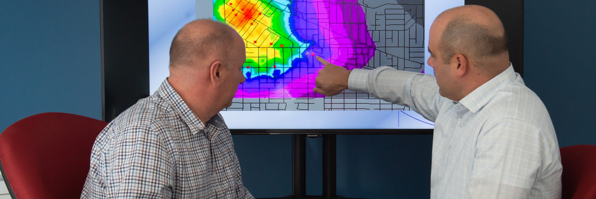 Two men sit while one points to a multi-coloured heat map on a screen.