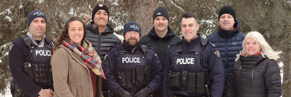Eight RCMP employees, some wearing police uniforms, stand outdoors in the snow smiling.