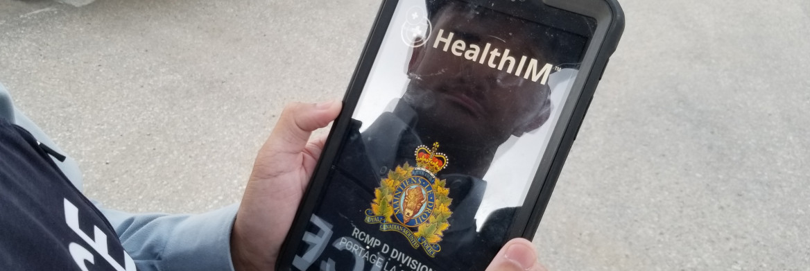 A male RCMP officer holding a tablet outdoors with his reflection visible on the screen.