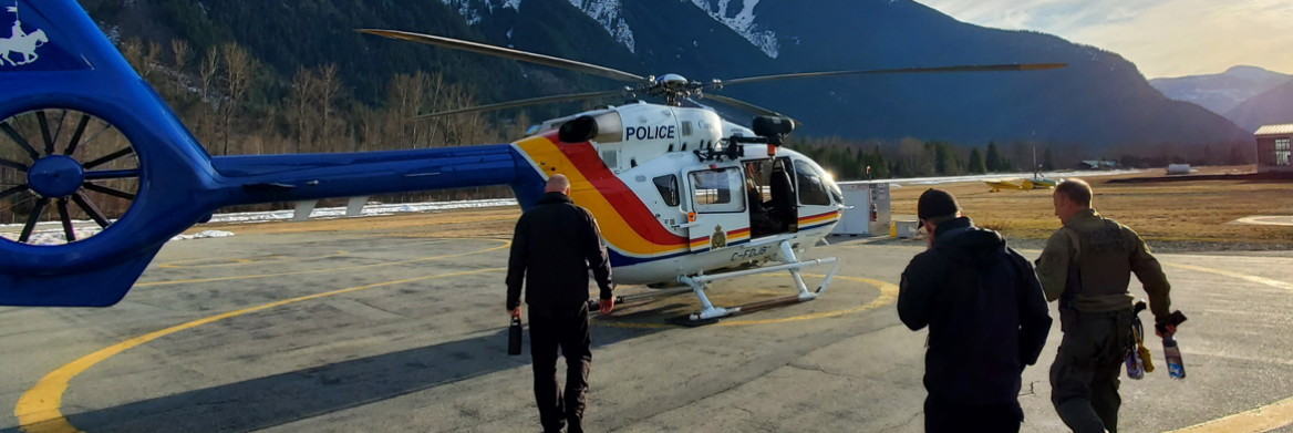 Two people in police uniforms kneel down in a in an equipped helicopter. A snow-covered mountain can be seen from the windows of the helicopter. 
