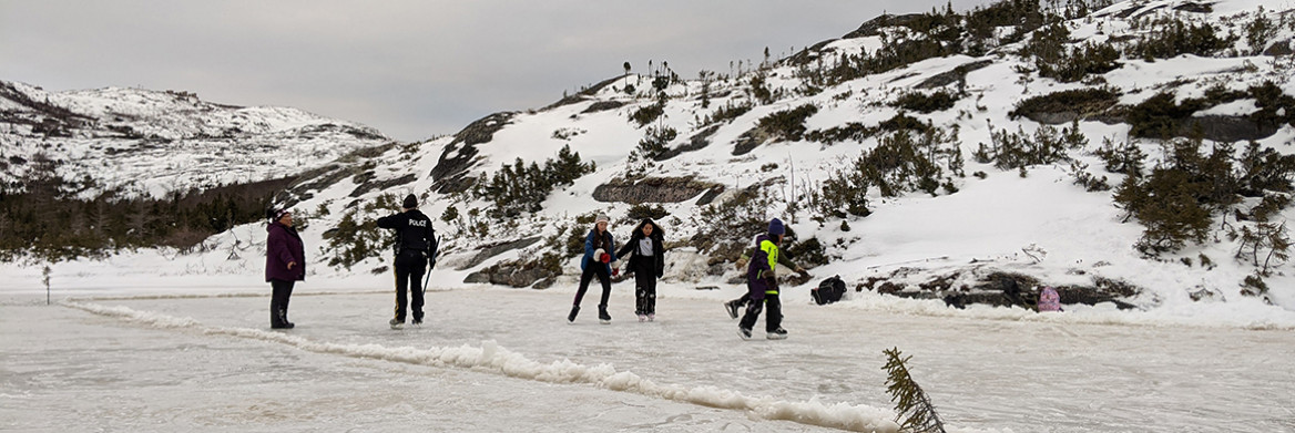 Un membre de la GRC, un autre homme et plusieurs jeunes patinant sur une patinoire extérieure entourée de collines enneigées et de conifères.