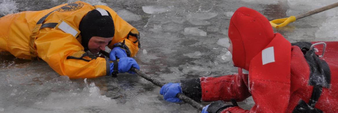 A man lying on a frozen lake uses a pole to help pull another man out of the ice. 