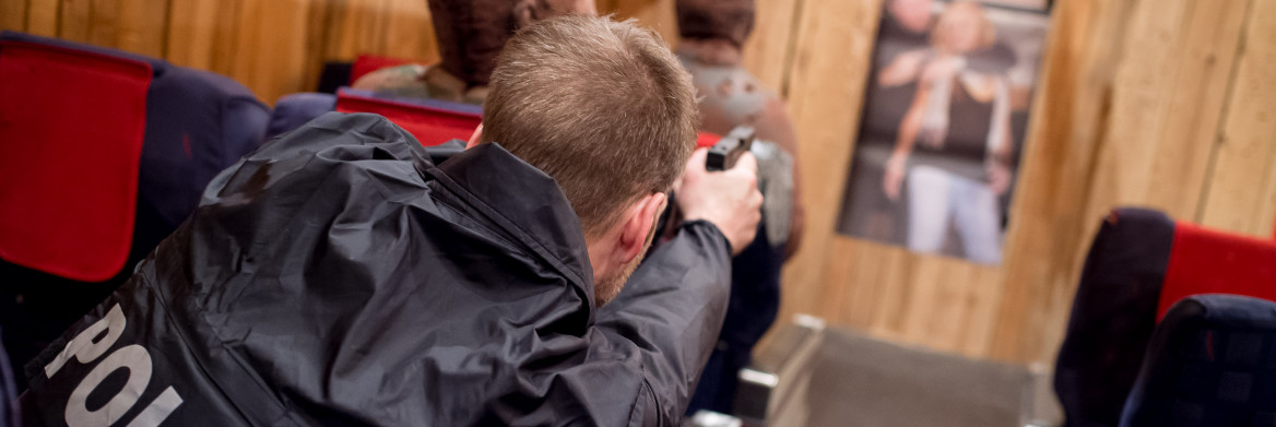 Man wearing a police jacket aims his gun at a target in an airplane aisle. 
