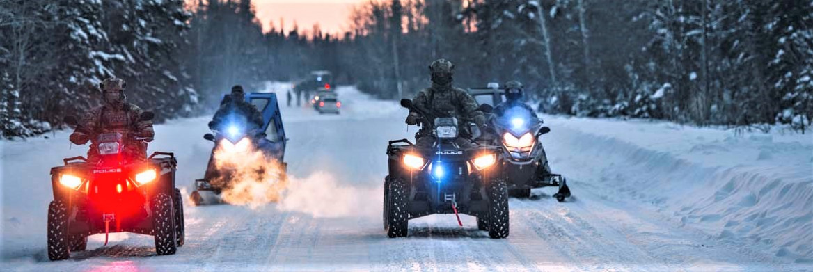At dusk, four people ride two all-terrain vehicles and two snowmobiles along a snow-covered road surrounded by coniferous trees. There are other people and vehicles in the distance.