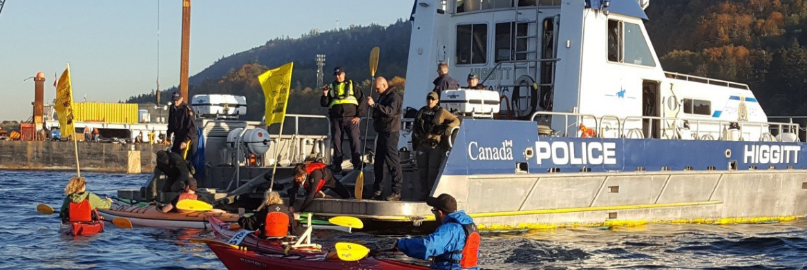 Four RCMP officers on vessel talk to four people in kayaks in open water.