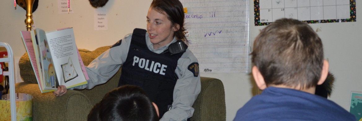 A female RCMP officer sits in a classroom while reading a book that she is showing to the students. A few young students sit in front of her.