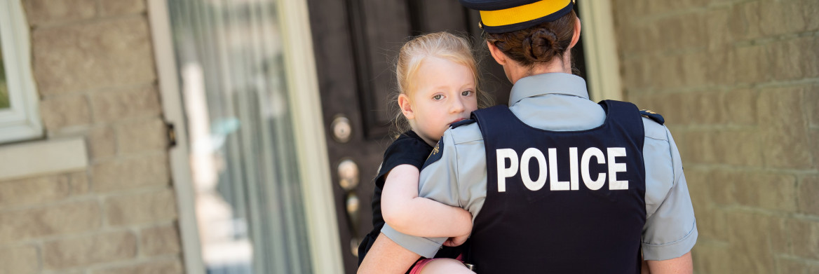 Femme policière tenant une fillette dans ses bras, devant la porte d'entrée d'un domicile.