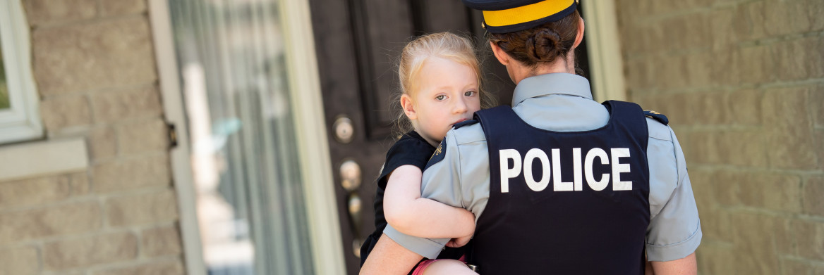 A female police officer holds a young girl in her arms standing at the front door of a house.
