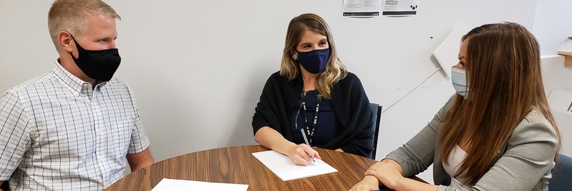 Un homme et deux femmes portant des masques, assis autour d'une table.