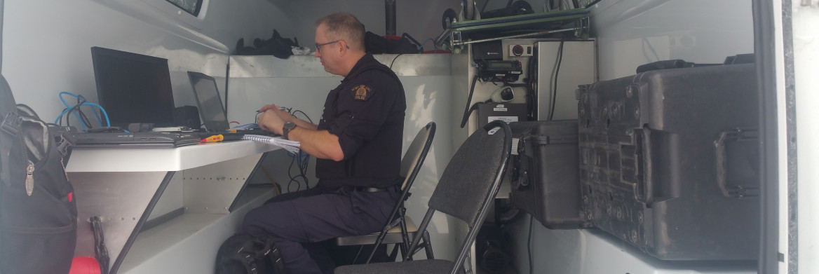 Male police officer sits at desk with computer in the back of a trailer.
