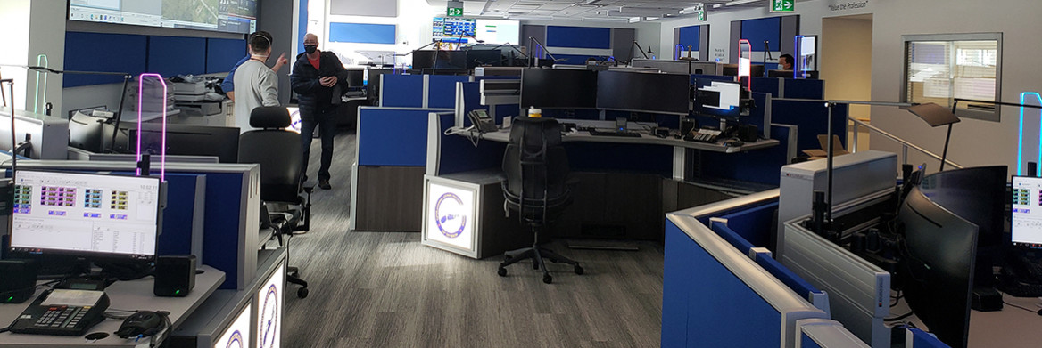 A room filled with state-of-the-art computer workstations. A group of men stand talking to each other as another male telecommunications officer sits at a computer to the right.