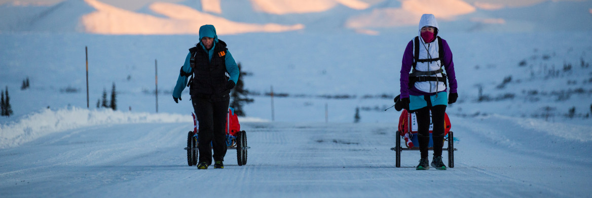 Deux femmes en habit de neige marchent le long d'une route glacée et enneigée avec des montagnes en arrière-plan.