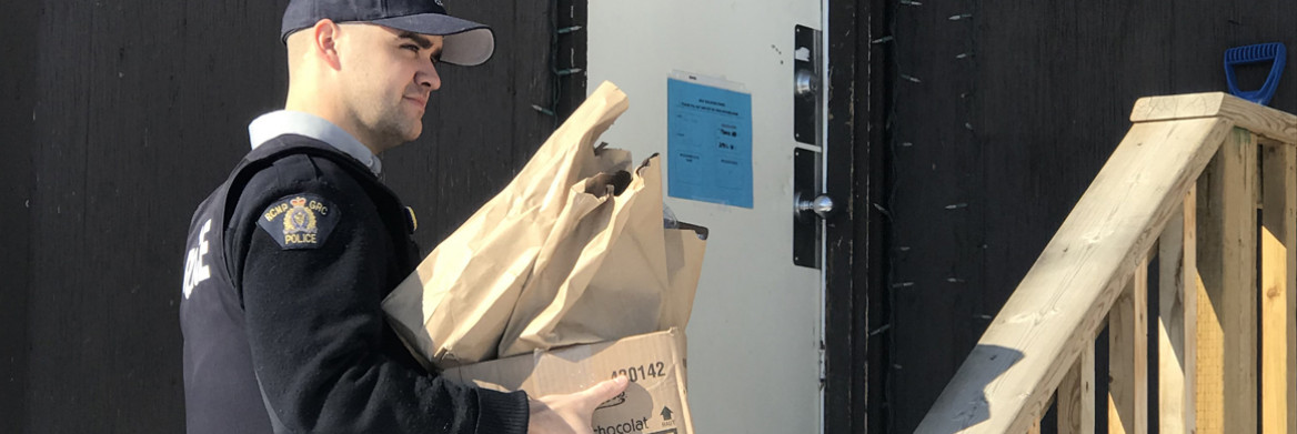 A male RCMP officer carries multiple brown bags and a box to a home. 