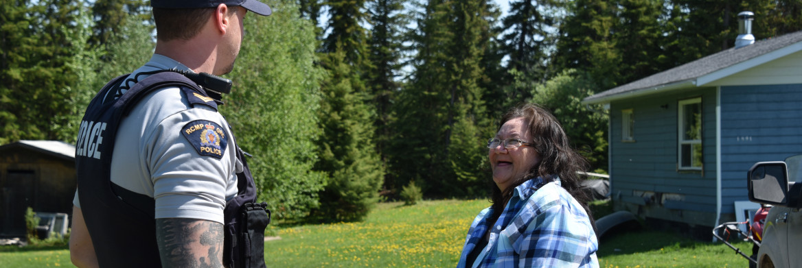 A male RCMP officer and an Indigenous woman face each other smiling in a treed yard next to a small blue house.