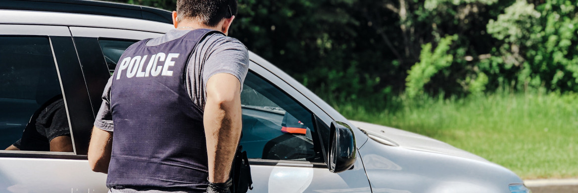 A male police officer in plainclothes stands near a vehicle outdoors.