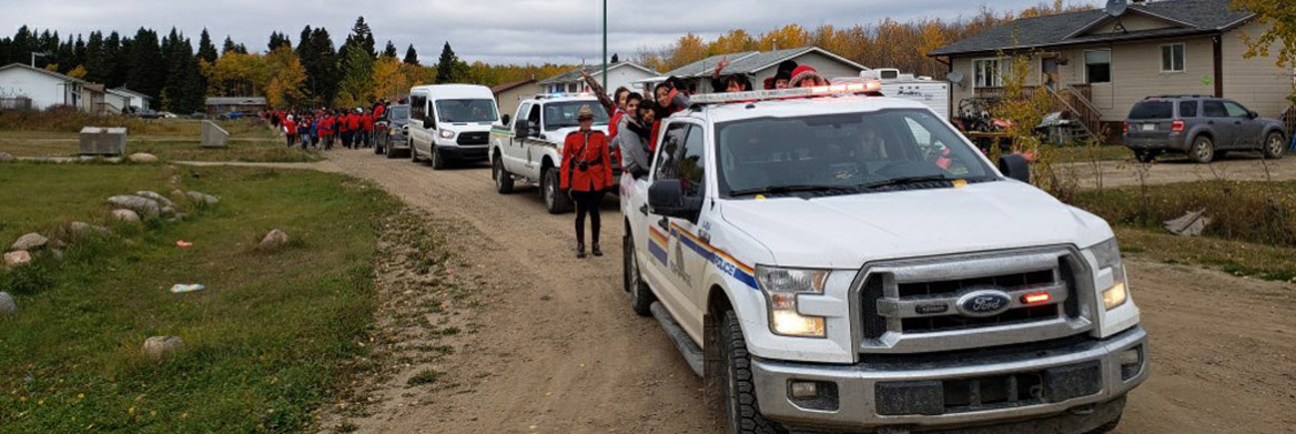 Une policière de la GRC en tunique rouge se tient debout à côté d'une camionnette de la GRC à l'arrière de laquelle se trouvent des gens qui agitent la main. Sur la route derrière eux se trouvent d'autres véhicules et une grosse foule de gens portant du rouge. 