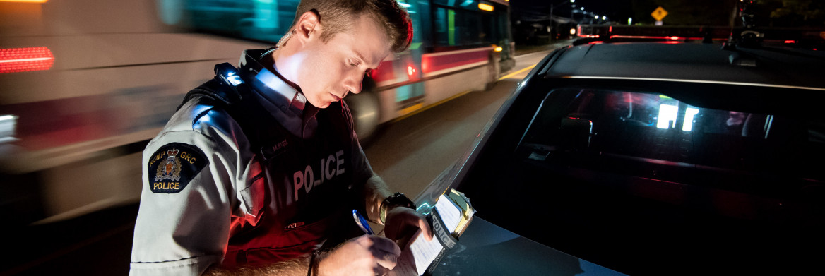 La nuit, un policier debout à côté de son auto-patrouille écrit dans son calepin, alors qu'un autobus passe derrière lui, dans le flou de l'arrière-plan. 