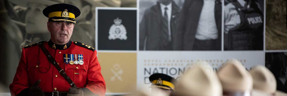 An RCMP officer in red serge faces a room of other RCMP officers in red serge wearing Stetsons. The wall behind him displays posters of police officers.