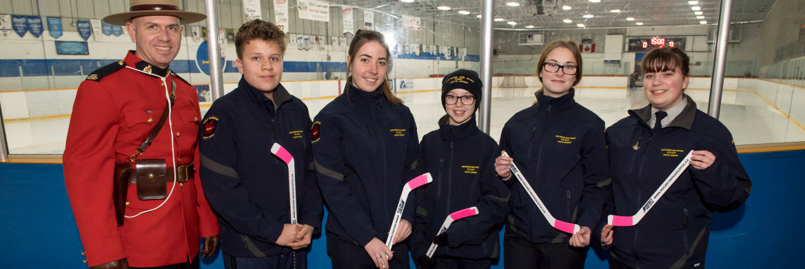 A male RCMP officer in red serge stands next to five youth holding pink-taped miniature hockey sticks. 