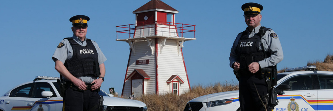 Two male RCMP officers stand two metres apart in front of a light house. Two police cruisers are in the background.