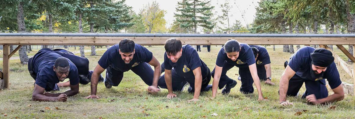 A diverse group of male and female pre-cadets crawl on a grassy field under a wooden beam in an outdoor obstacle course. 