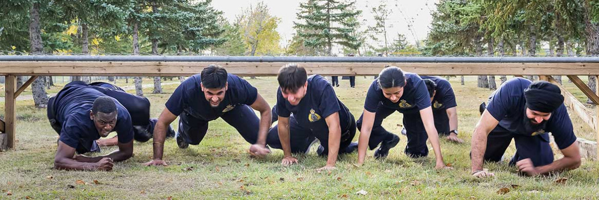 A diverse group of male and female pre-cadets crawl on a grassy field under a wooden beam in an outdoor obstacle course.