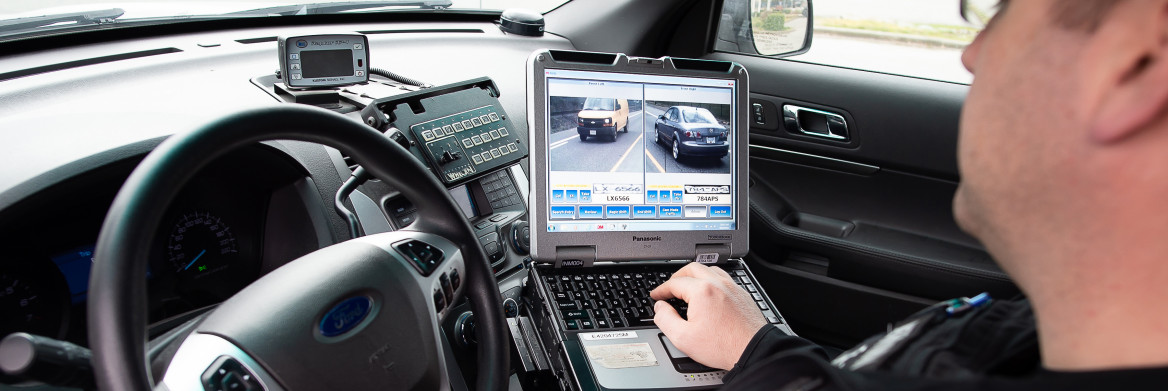 Male police officer sits in police car looking at computer screen.