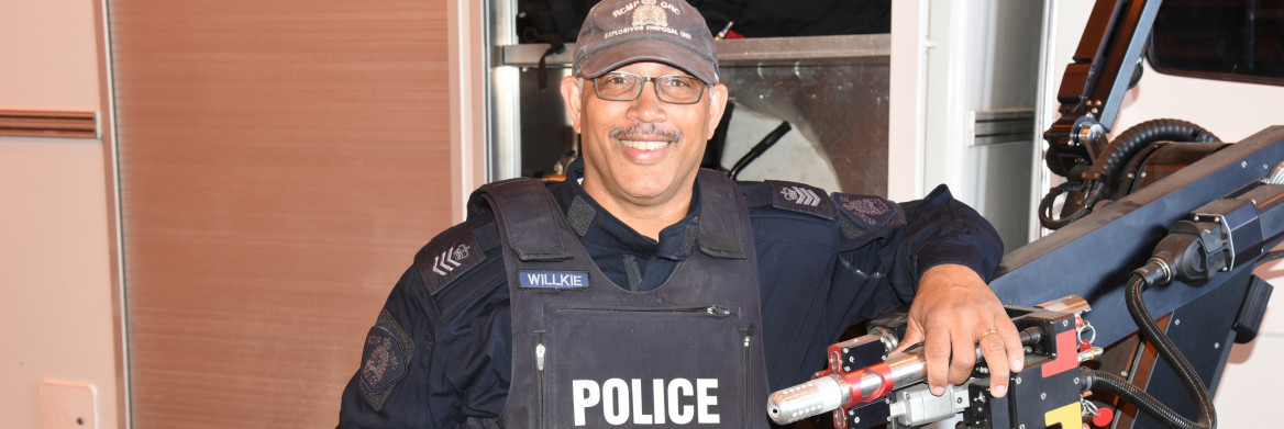 A male RCMP officer stands in front of a large water cannon.