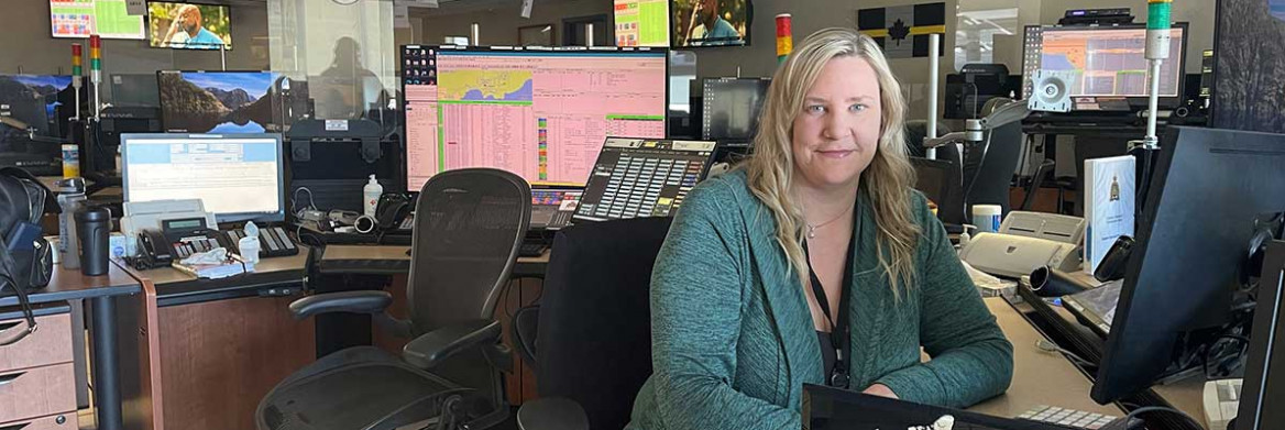 A woman, wearing a headset, sits at a desk in front of computer screens.