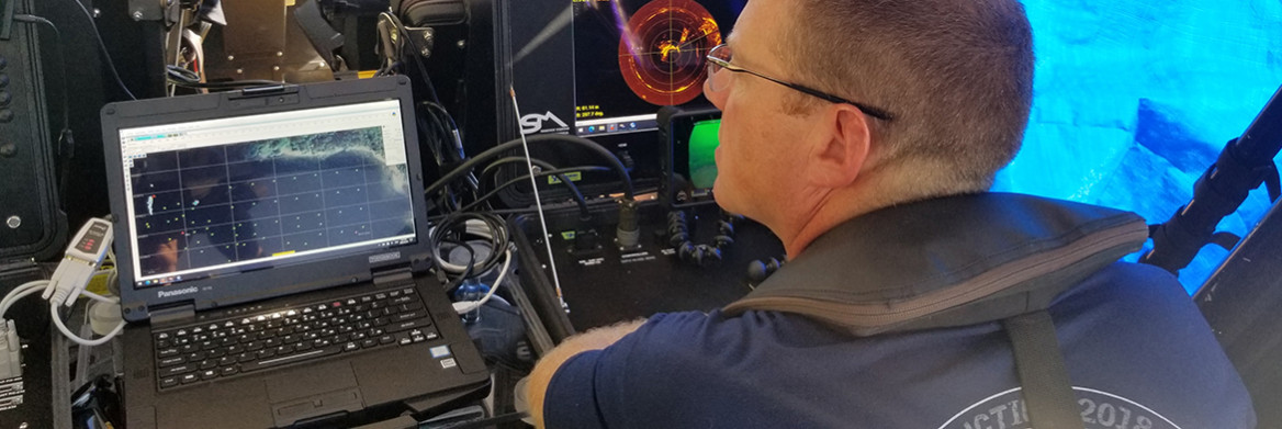 A male RCMP officer looks at two screens that display shapes from the sea floor.