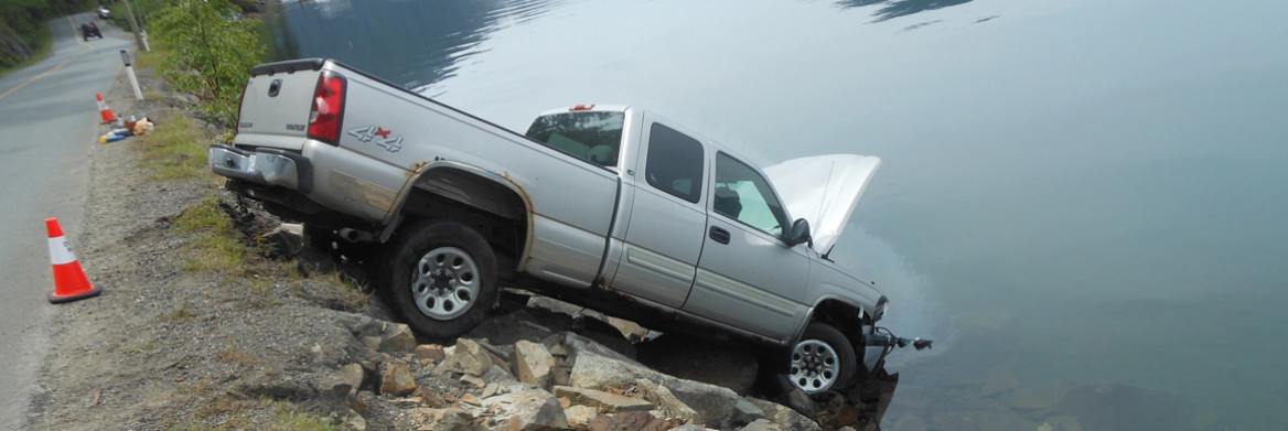 Une camionnette sur un talus rocailleux dont le capot moteur est partiellement submergé dans un lac. En arrière-plan, des conifères et le relief escarpé d'une montagne.