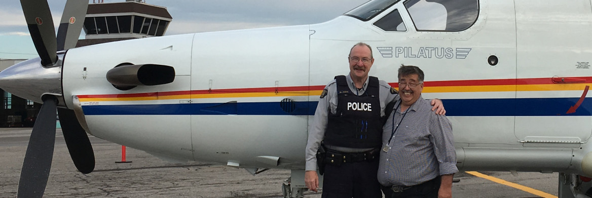 One male police officer and another man stand arm in arm and smiling in front of an airplane. 