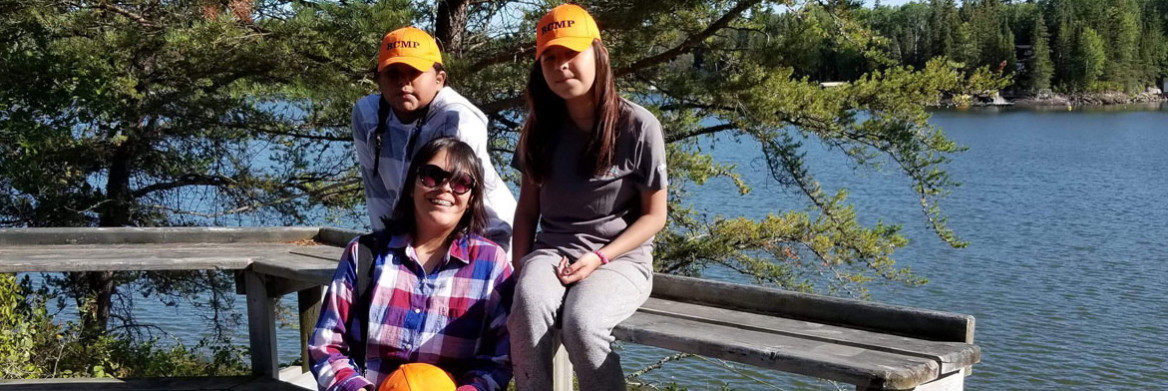 Two girls wearing orange caps sit in front of a lake with a woman. 