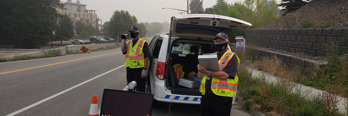 Deux hommes vêtus de gilets de haute visibilité debout près d'une route avec un radar de vitesse. 