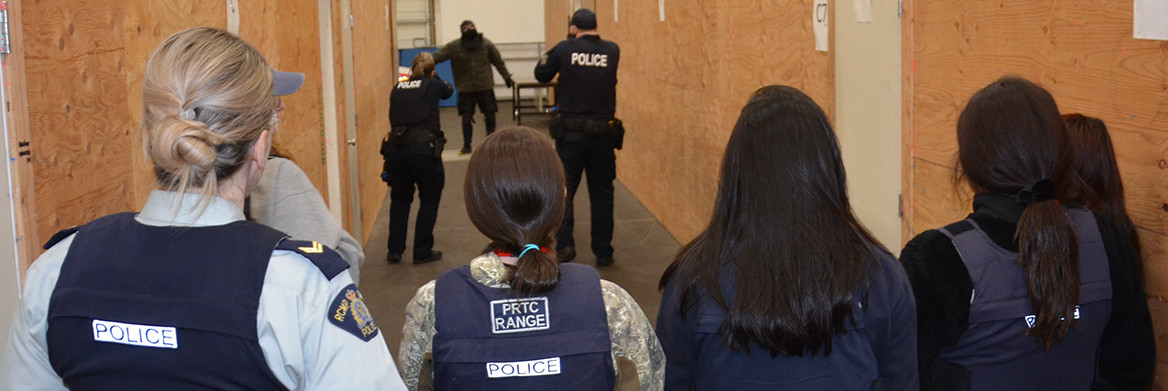 A female police officer and several young people look down a hallway toward two police officers (1 male, 1 female) with their pistols drawn as they approach a male actor in plainclothes wearing a bandana.