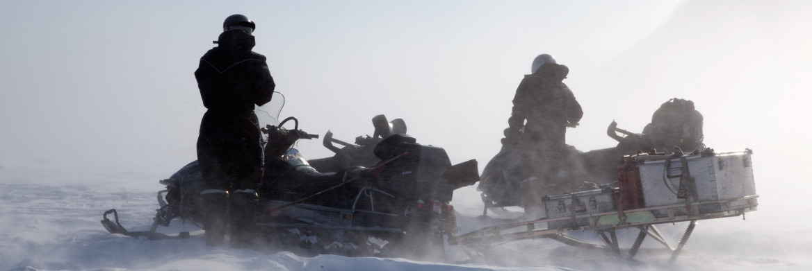Two snowmobilers surrounded by blowing snow on barren landscape.