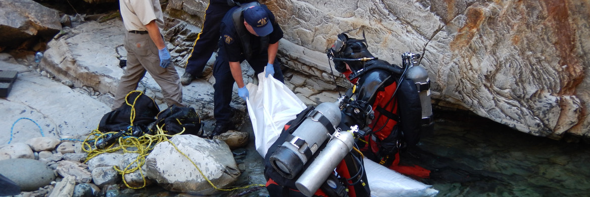Two RCMP divers pull a bagged drowning victim from the water while male officers retrieve it from a rocky shore.