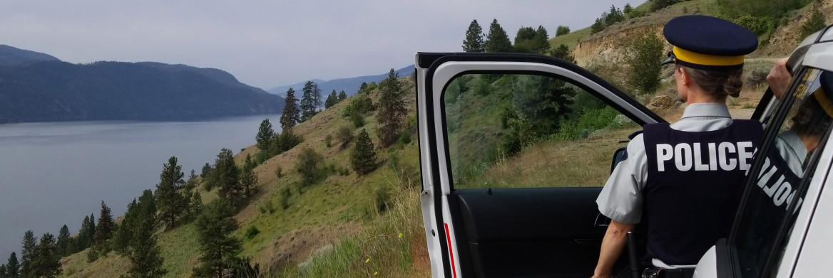 A female RCMP officer stands beside a vehicle, with an open door, facing a lake.