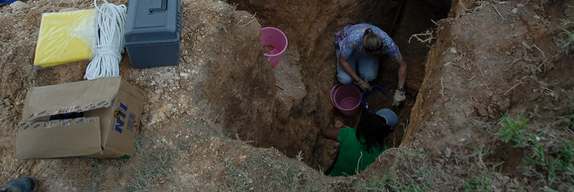 Two women collect evidence from a deep hole in the ground.
